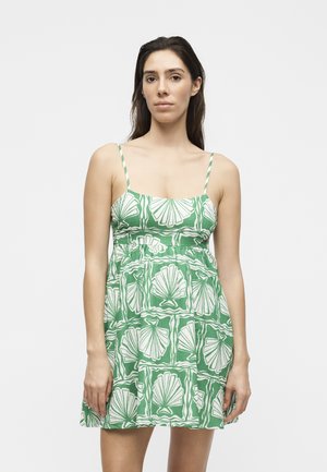 Γυναίκα με μακριά σκούρα μαλλιά φοράει πράσινο φόρεμα με λεπτές τιράντες και λευκά σχέδια με κοχύλια, στέκεται μπροστά σε ένα απλό ανοιχτό φόντο.