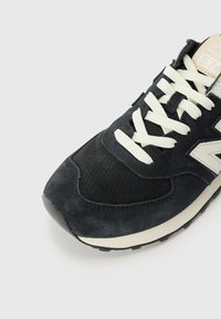 Baskets en suède noir avec une boîte à orteils perforée, lacets crème et semelle intermédiaire blanche. Présente un logo "N" gris proéminent sur le côté.