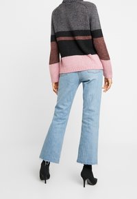 Sweter z dzianiny z wysokim kołnierzem, z poziomymi paskami w odcieniach szarości, czerni, terakoty i różu; w połączeniu z jasnoniebieskimi spodniami dzwonami.