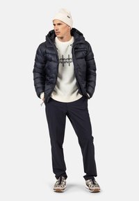 Giubbotto imbottito blu navy, materiale leggero; maglione crema con design grafico; pantaloni scuri; berretto lavorato a maglia crema; scarpe in tonalità neutra con accenti.