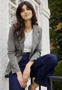 Młoda kobieta z długimi ciemnymi włosami, ubrana w szarą marynarkę, biały top i niebieskie dżinsy, siedzi na zewnątrz na poręczy obok białego budynku.