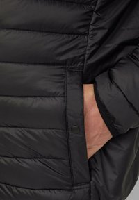 Zwarte gewatteerde jas met horizontale stiknaad, zijzak met drukknop en elastische manchetten. Soepele, lichte stoftextuur.