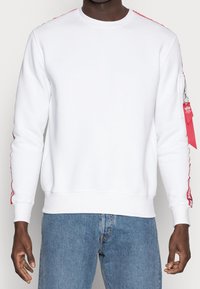 Uomo che indossa una felpa bianca con striscia rossa e testo sulle maniche, tasca con zip rossa sul braccio sinistro, abbinata a jeans blu.