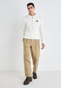 White ribbed zip-up jacket with a wide collar, paired with tan pleated trousers and brown lace-up boots. Features a small logo on the jacket.