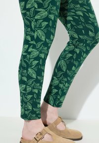 Leggings verts avec un motif de feuilles en vert clair. Le matériau semble doux avec un design ajusté. Chaussures claires avec un détail de boucle.