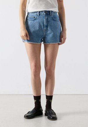 Person trägt hellblaue Jeansshorts, weißes T-Shirt, schwarze Socken mit Text und schwarze Loafer und steht auf einem schlichten hellen Boden.