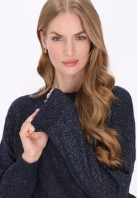 Pulover tricotat bleumarin cu un finisaj texturat, având accente strălucitoare. Manșetele ribbate și o croială lejeră conturează decolteul și umerii.