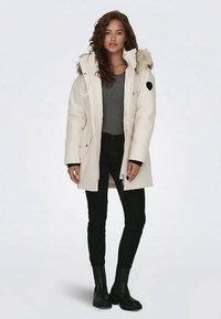 Γυναίκα φορώντας ένα άσπρο χειμερινό παλτό με κουκούλα από γούνα, γκρι πουκάμισο, μαύρα παντελόνια και μαύρες μπότες, στέκεται μπροστά σε ένα απλό φωτεινό φόντο.