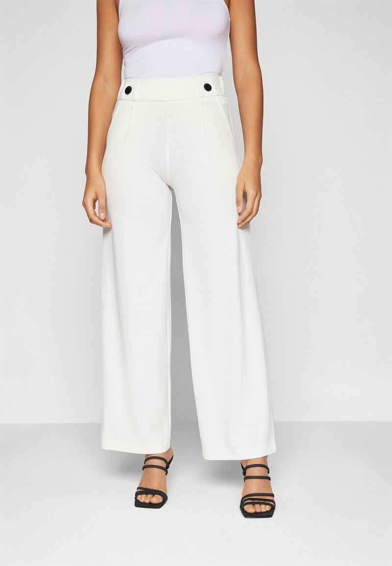 Pantaloni bianchi a gamba larga con una texture liscia, vita alta, doppie tasche frontali e dettagli in bottone nero, abbinati a sandali neri con strappi.