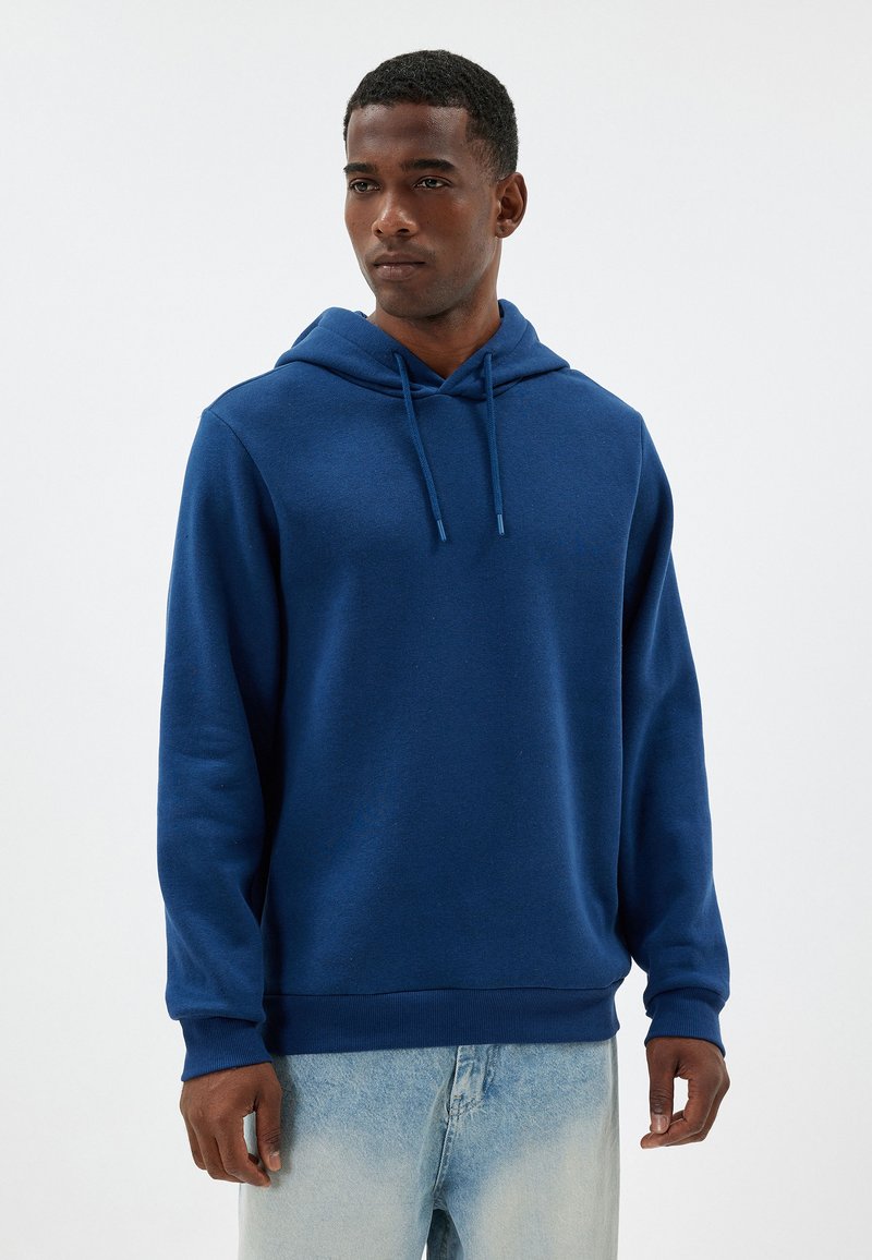 Sudadera azul con un bolsillo canguro delantero, capucha ajustable con cordón y puños acanalados. Combinada con jeans claros desgastados. Textura suave.