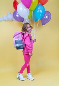 Rosa Rucksack mit grauen Akzenten, verstellbaren Trageriemen und Fronttasche. Kind in rosa Outfit hält bunte Ballons vor einem gelben Hintergrund.