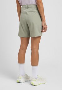 Helle grüne Shorts mit glatter Textur, Bund mit Gürtelschlaufen und einem Logo. Tragen Sie dazu weiße Socken und hellfarbene Turnschuhe.