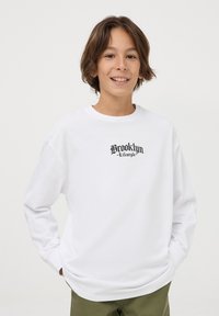 T-shirt bianca a maniche lunghe in cotone con scollo rotondo, caratterizzata da una grafica nera "Brooklyn Lifestyle" sul petto.