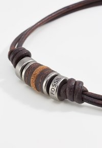 Brown leather necklace featuring metal and suede accents with a round metallic centerpiece labeled "FOSSIL." Textured and braided design.