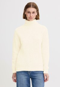 Krämfärgad polotröja med ribbad textur, avslappnad passform och långa ärmar, i kombination med blå jeans.