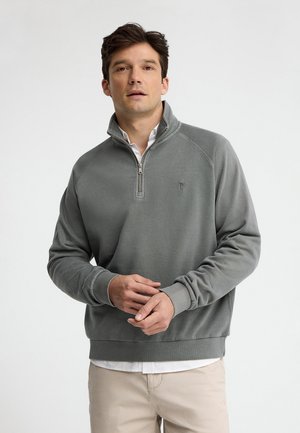 Uomo con pullover grigio con zip a quarto e pantaloni beige, che sistema i polsini, rivolto frontalmente su sfondo bianco uniforme.