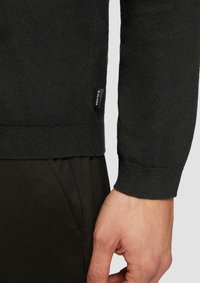 Donkergroen trui met geribde manchetten en zoom, voorzien van een klein zwart label met "SOLIVER". Zachte textuur, gedeeltelijk zichtbare hand en broek.