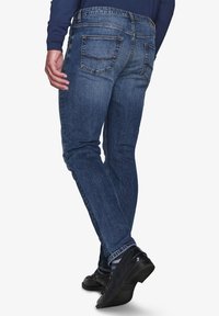 Jeans in denim di colore blu scuro, dalla forma aderente. Le caratteristiche includono tasche posteriori, leggera sbiaditura sulle cosce e cuciture a contrasto. Indossati con scarpe nere.