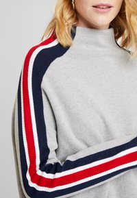 Szary sweter z wysokim kołnierzem, ozdobiony prążkowanymi akcentami w kolorze granatowym i czerwonym na rękawach. Miękka tekstura, luźny krój i minimalistyczny design.