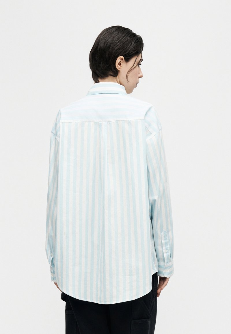 Persona con capelli corti e scuri, vista di lato, che indossa una camicia a maniche lunghe, larga, a righe verticali azzurre e bianche e pantaloni scuri.