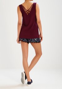 Mörk vinröd linne med korsade ryggband, tillsammans med svarta shorts med färgglada blommönster och spetsdetaljer, bärs med marinblå sneakers.