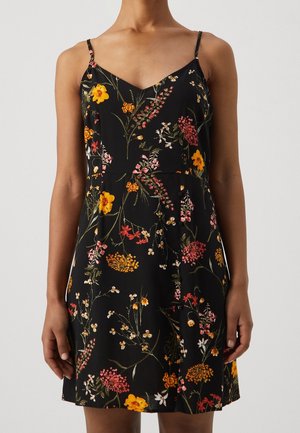Vestido negro sin mangas con tirantes finos y estampado floral multicolor, llevado por una persona que está de pie frente a un fondo liso.