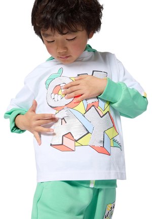 Copil atingând litere cu paiete care formează cuvântul „JOACĂ” pe un hanorac colorat, purtând mâneci și pantaloni verzi asortați.