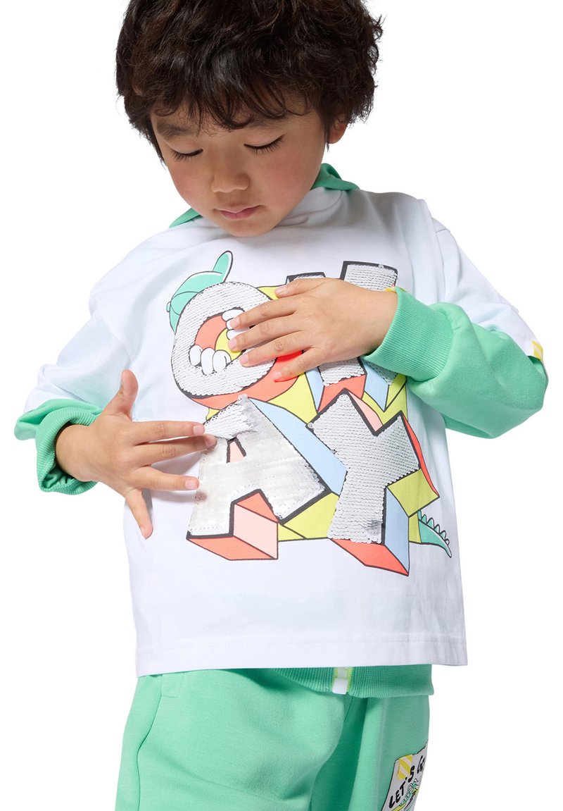 Dziecko dotyka cekinowych liter tworzących napis „PLAY” na kolorowej bluzie, nosząc dopasowane zielone rękawy i spodnie.