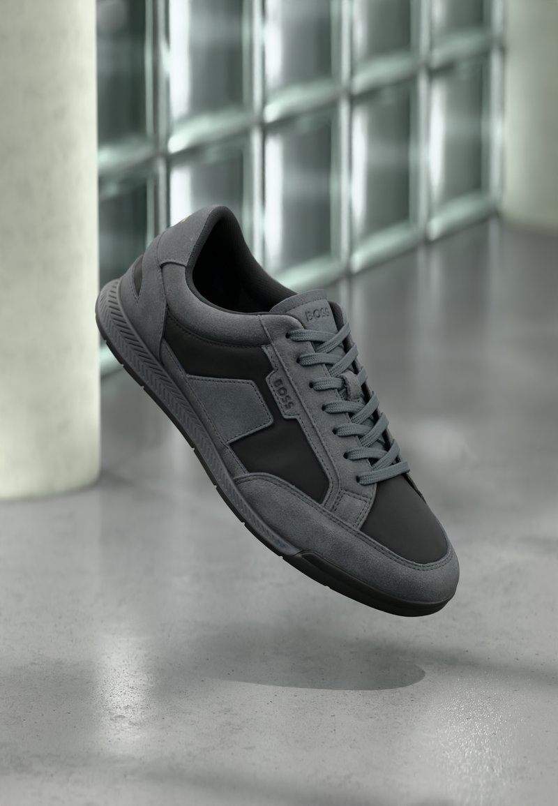 Chaussure de sport grise combinant suède et matériaux synthétiques, avec un laçage et une semelle texturée pour une meilleure adhérence.