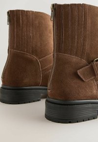 Stivaletti in camoscio marrone con zip sul retro, dettagli cuciti, accenti a strappo e suole in gomma nera spessa su uno sfondo neutro.