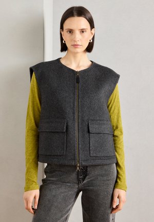Grijze wollen mouwloze vest met een ronde halslijn, een rits aan de voorkant en twee voorzakken, gedragen over een langmouwige gele top.