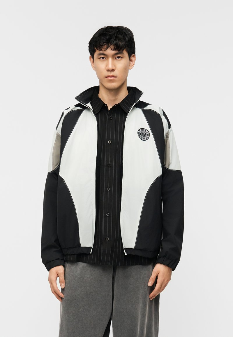 Νέος άνδρας που φοράει μαύρο και άσπρο ζακέτα με φερμουάρ πάνω από μαύρο πουκάμισο με ρίγες και γκρι παντελόνι, στέκεται μπροστά σε απλό φόντο.