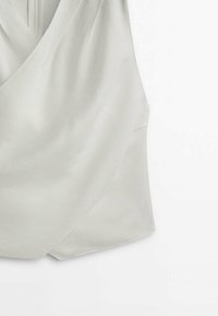 Haut sans manches beige clair avec un devant style cache-cœur et un tissu subtilement texturé sur fond blanc.