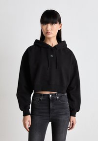 Black cropped hoodie with a drawstring hood and a small logo patch. Paired with high-waisted black jeans featuring front pockets.