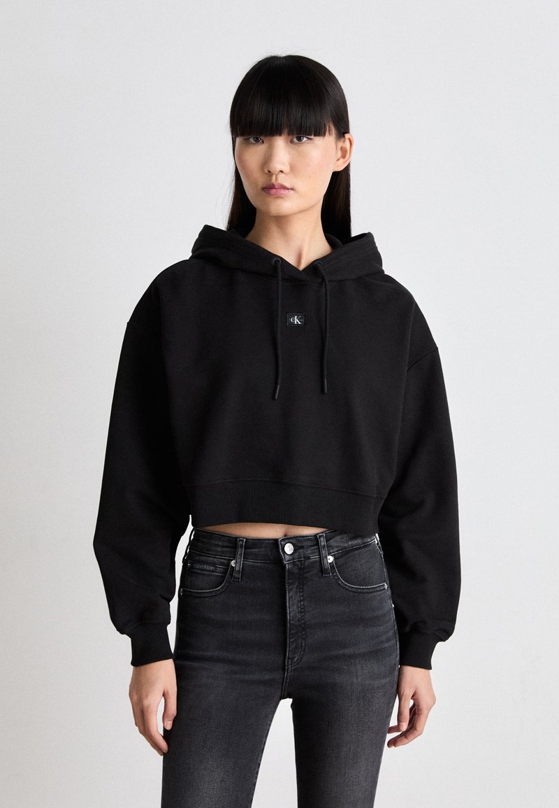 Black cropped hoodie with a drawstring hood and a small logo patch. Paired with high-waisted black jeans featuring front pockets.