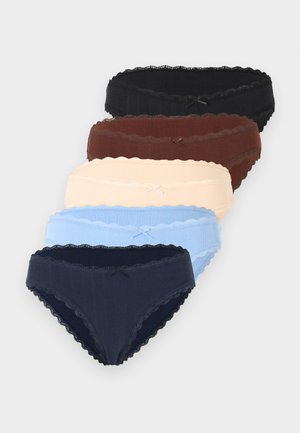 Femme aux longs cheveux ondulés portant un soutien-gorge beige et une culotte assortie bordée de dentelle, debout avec les mains derrière le dos sur un fond uni.