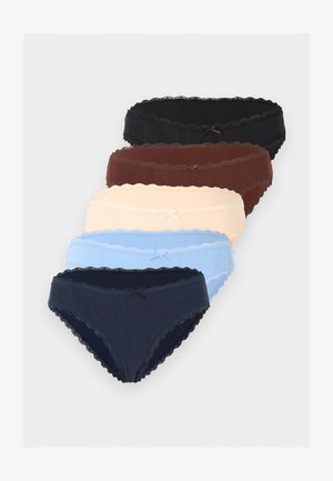 Hosszú, hullámos hajú nő, aki bézs melltartót és hozzáillő, csipkével szegélyezett fehérneműt visel, háttal a kezét a hát mögött tartva egyszínű háttér előtt áll.