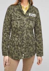 Groene jas met camouflagepatroon, gemaakt van katoen; voorzien van twee borstzakken, knoopsluiting en een patch met de tekst "Dromers."