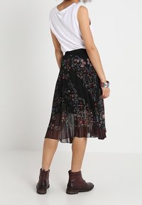 Jupe plissée noire avec broderies florales colorées, associée à un haut sans manches blanc et des bottines à lacets marron.