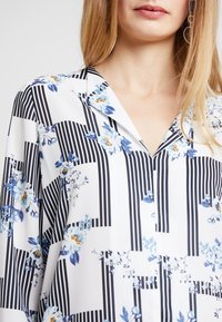 Camicia bianca con strisce verticali nere e motivi floreali in blu e giallo. Tessuto morbido con colletto e vestibilità ampia.