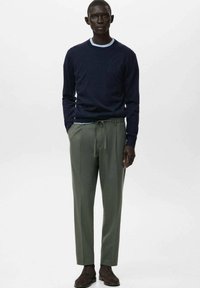 Hombre con suéter azul marino, camiseta interior azul claro, pantalones verdes oliva con cordón y zapatos de ante marrón, de pie con las manos parcialmente en los bolsillos.