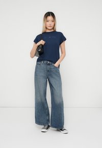 Junge Frau trägt ein dunkelblaues T-Shirt, weite, ausgebleichte blaue Jeans, schwarze Sneakers, hält eine schwarze Handtasche über der Schulter und steht vor einem weißen Hintergrund.