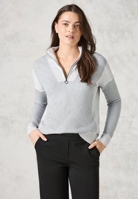 Grauer und hellgrauer Quarter-Zip-Pullover mit geripptem Kragen und Bündchen, aus weichem Stoff gefertigt. Kombiniert mit schwarzen Hosen, die Hände in den Taschen.