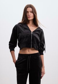 Sort cropped velour zip-up jakke med lange ærmer, frontlomme og snøre-detalje. Glat tekstur, parret med sorte velourbukser.