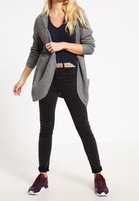 Szary dziergany kardigan z otwartym przodem, czarne dopasowane dżinsy i bordowe sneakersy. Gładka faktura, luźne dopasowanie oraz głęboki dekolt w kształcie litery V pod spodem.