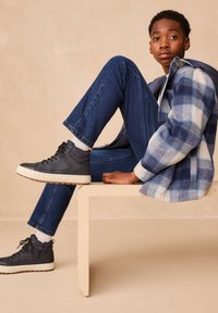 Giacca in flanella a quadri blu e bianca, jeans blu scuro, sneaker alte grigie con suole bianche, seduto su una panchina di colore chiaro.