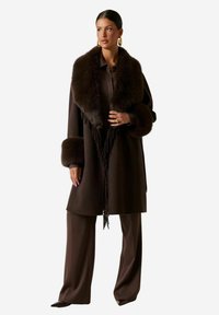 Cappotto marrone con ampio colletto e polsini in pelliccia, con frange lungo la parte frontale. Indossato sopra pantaloni marroni abbinati.