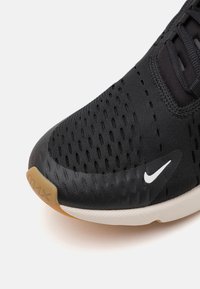 Fekete sportcipő, textúrált hálós felsőrésszel és fehér Nike logóval. Párnázott talppal rendelkezik, gumitalppal és világos barna kiegészítőkkel.