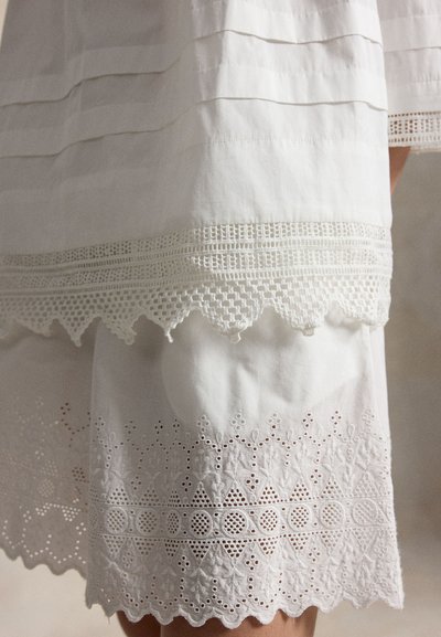 Λευκό ύφασμα με στρωμένο δαντέλα και κέντημα eyelet με κυματιστές άκρες σε ένα ένδυμα, πάνω σε ουδέτερο φόντο.