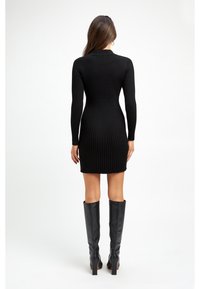 Vestito nero in maglia a coste con maniche lunghe, silhouette aderente e lunghezza a metà coscia. Abbinato a stivali neri in pelle al ginocchio.